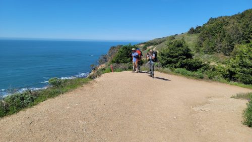 Two hikers walk along a dirt path by the ocean, surrounded by greenery and a clear blue sky.