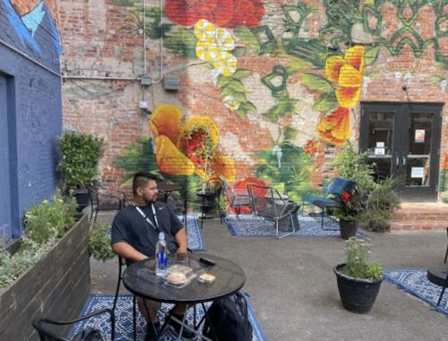 A man sits at a table in a vibrant outdoor space with a colorful mural and potted plants around him.