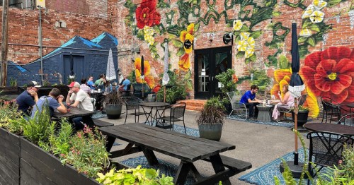 A vibrant outdoor café with colorful murals, featuring people seated at tables among plants and flowers.