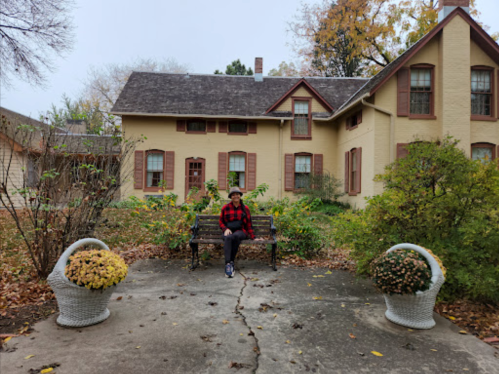 A person in a red plaid shirt sits on a bench in front of a large, yellow house surrounded by autumn foliage.
