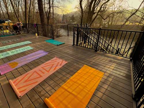 A peaceful outdoor yoga space with colorful mats arranged on a wooden deck overlooking a river and trees.