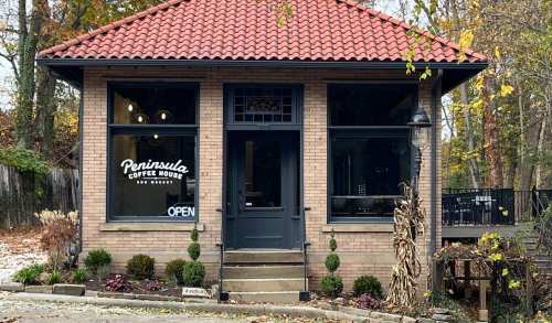 A cozy brick coffee house with a tiled roof, large windows, and a welcoming "Open" sign. Surrounded by autumn foliage.