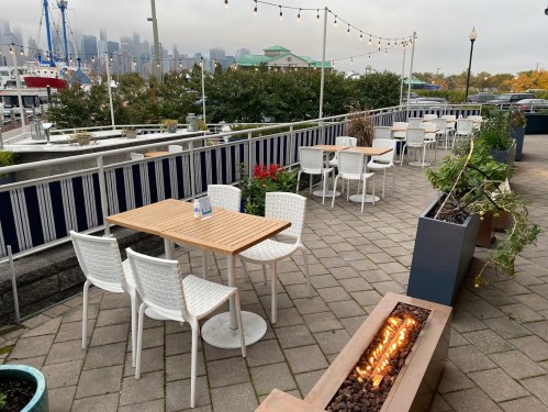 Outdoor dining area with wooden tables, white chairs, and a fire pit, overlooking a marina and city skyline.