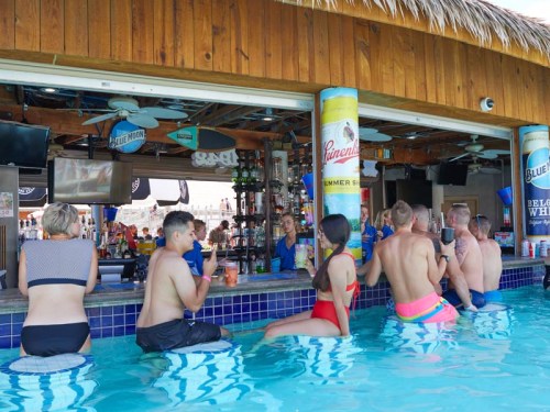A lively poolside bar scene with people enjoying drinks, some seated in the water, under a thatched roof.