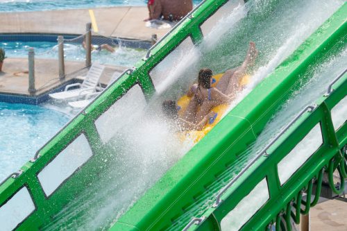 Two people slide down a green water slide, splashing into a pool below, with others enjoying the water nearby.