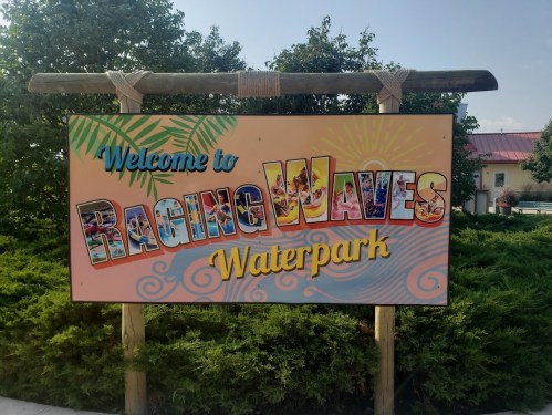 Colorful sign reading "Welcome to Raging Waves Waterpark" surrounded by greenery and a clear blue sky.