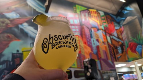 A hand holds a cocktail glass with a lime wedge in front of a colorful mural at Obscurity Brewing & Craft Mead.