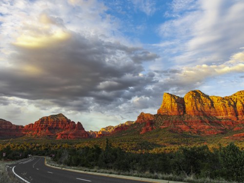 Scenic view of red rock formations under a cloudy sky, with a winding road in the foreground.