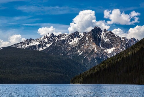 A serene lake reflects towering snow-capped mountains under a blue sky with fluffy clouds. Lush green forests line the shore.