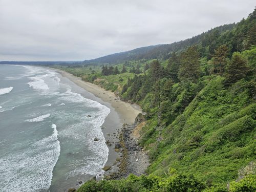 A scenic coastal view featuring a sandy beach, rocky shoreline, and lush green hills under a cloudy sky.