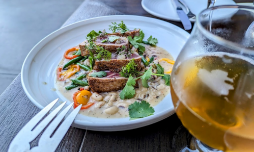 A plate of sliced breaded chicken with vegetables in a creamy sauce, alongside a glass of beer on a wooden table.