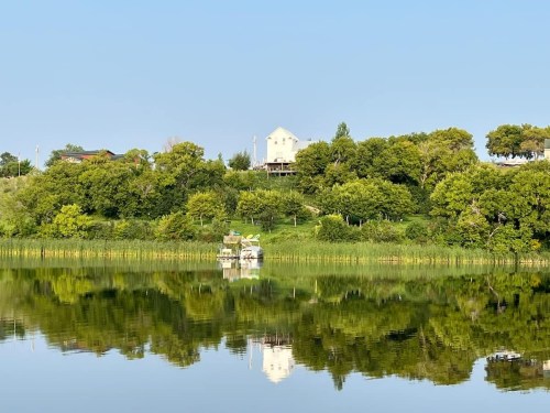 A serene lake reflects a green hillside with a white house and trees under a clear blue sky.