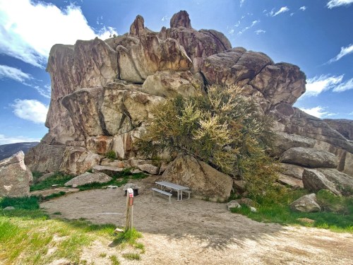 A large rock formation with a tree and a picnic table in a grassy area under a bright blue sky.
