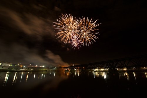 Colorful fireworks burst over a river at night, reflecting light on the water and illuminating a bridge in the foreground.