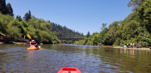 A kayaker paddles on a calm river surrounded by lush greenery, with a bridge and people on the shore in the background.