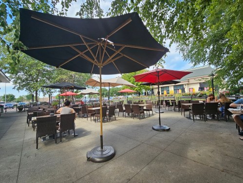 Outdoor dining area with large umbrellas, tables, and chairs surrounded by trees and a view of the water.