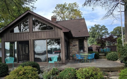 A cozy brown house with large windows, surrounded by greenery and outdoor seating on a wooden deck.