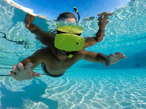 A person swimming underwater in a pool, wearing a yellow snorkeling mask and reaching out with their hands.