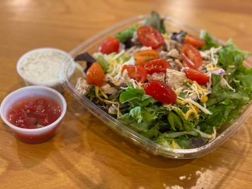 A fresh salad with greens, cherry tomatoes, shredded cheese, and grilled chicken, served with ranch and salsa on the side.