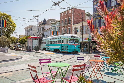 A vintage tram passes through a colorful street with outdoor seating and vibrant buildings in a sunny urban setting.
