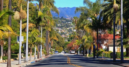An empty street in Santa Barbara, California, lined with palm trees and a hillside covered in homes in the distance.