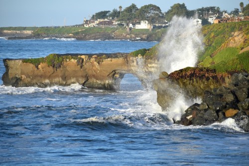 Waves crash against a rocky coastline with a natural arch, surrounded by lush greenery and distant houses.