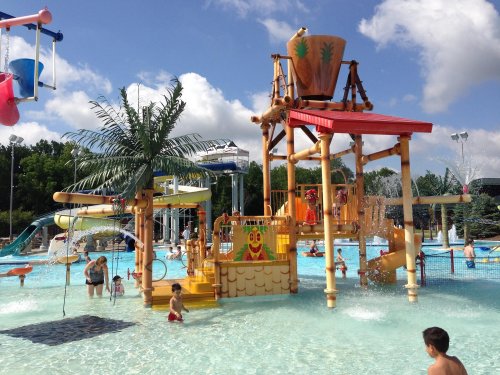 A vibrant water park scene with a large play structure, slides, and children enjoying the water under a blue sky.