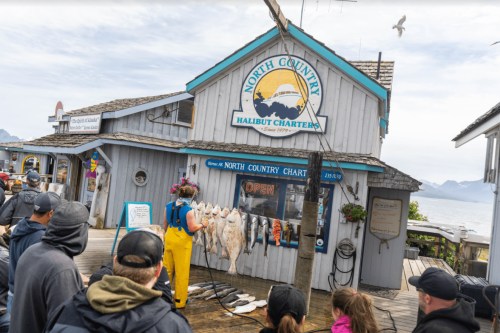 A woman in yellow overalls displays freshly caught halibut outside a fishing charter shop, with a crowd watching.