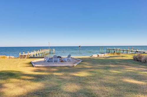 A serene waterfront scene featuring a grassy area, a fire pit, and a wooden pier extending into the calm blue water.