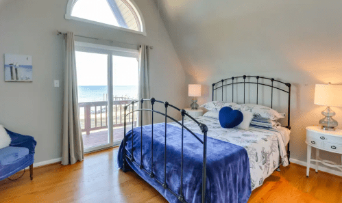 Cozy bedroom with a blue bedspread, large windows, and ocean view, featuring a chair and bedside lamps.