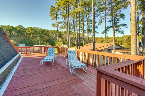 A wooden deck with two lounge chairs, surrounded by tall trees and rooftops in the background.