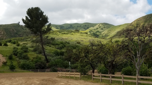 Lush green hills under a cloudy sky, with a dirt path and wooden fence in the foreground.