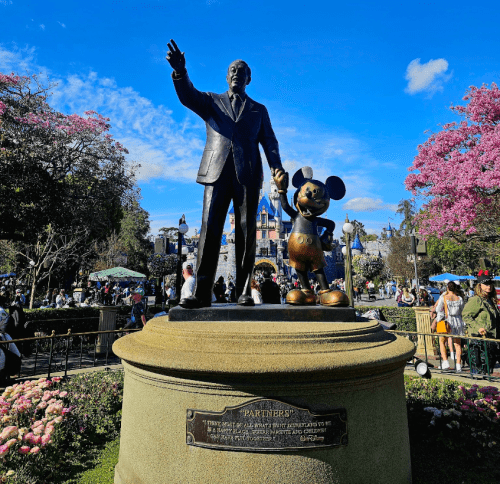 Statue of Walt Disney and Mickey Mouse in front of a castle, surrounded by blooming trees and a lively park atmosphere.