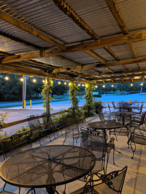Outdoor seating area with metal tables and chairs, string lights, and greenery under a wooden roof at dusk.