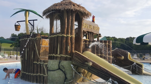 A wooden water play structure with slides and a palm tree, featuring a child playing nearby in a pool area.
