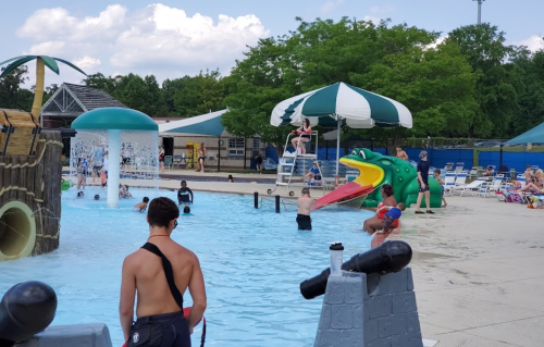 A lively pool scene with children playing, a large frog slide, and a lifeguard on duty under a shaded area.