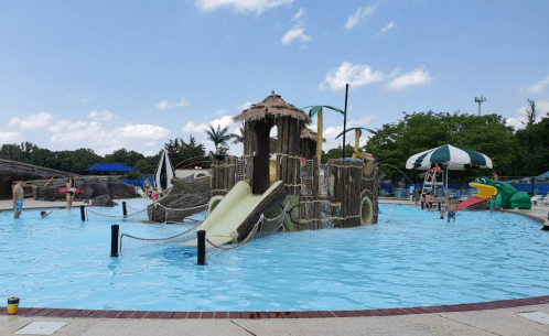 A water park with a large play structure, slides, and children swimming in a blue pool under a partly cloudy sky.