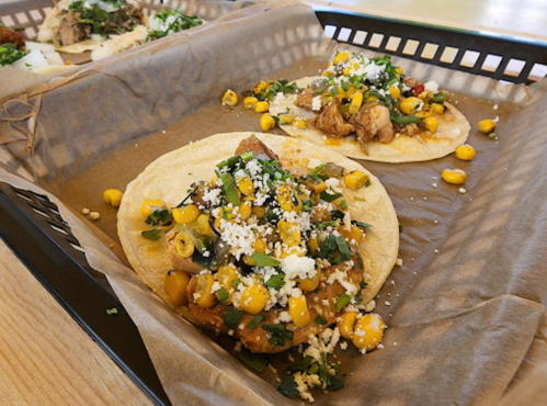 Two tacos on a tray, topped with chicken, corn, cheese, and fresh herbs, served on parchment paper.