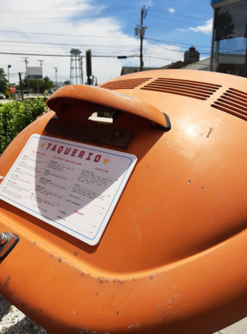 An orange taqueria sign with a menu, set against a blue sky and power lines in the background.