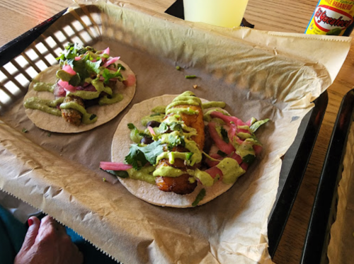 Two tacos on a tray, filled with fried fish, avocado sauce, pickled onions, and cilantro, served with a drink.