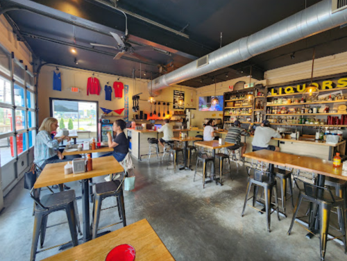 A casual bar interior with wooden tables, patrons seated, and a shelf stocked with liquor in the background.