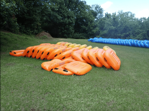 A row of orange and yellow inflatable tubes stacked on green grass, with trees in the background.