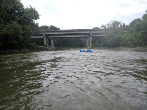 A blue raft floats on a river beneath a concrete bridge, surrounded by lush greenery and cloudy skies.