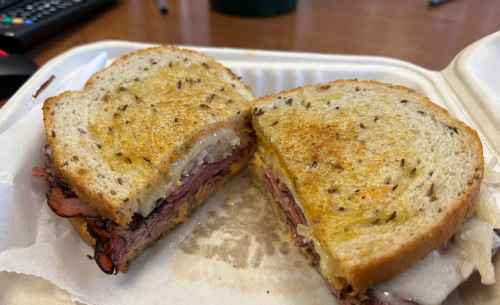 A close-up of a sliced sandwich on rye bread, filled with layers of meat and cheese, resting in a takeout container.