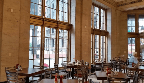 Interior of a cafe with large windows, wooden tables, and chairs, overlooking a city street.