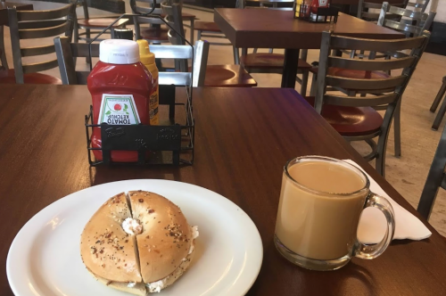 A bagel with cream cheese on a plate, accompanied by a cup of coffee, with ketchup and mustard on a table.