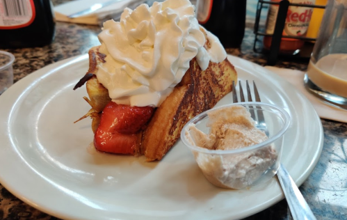 A slice of French toast topped with whipped cream and served with a side of cinnamon cream in a small cup.