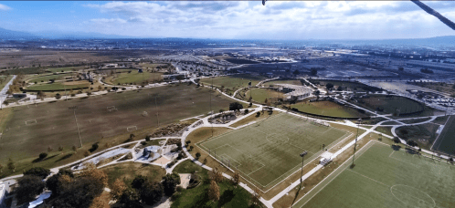 Aerial view of expansive sports fields and parks, with mountains and a cityscape in the background under a cloudy sky.