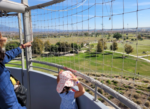 A child in a sun hat looks out from a tower, pointing at a park with sports fields and trees in the background.