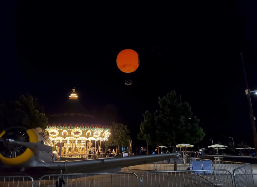A brightly lit carousel at night with a large orange balloon floating above and trees in the background.